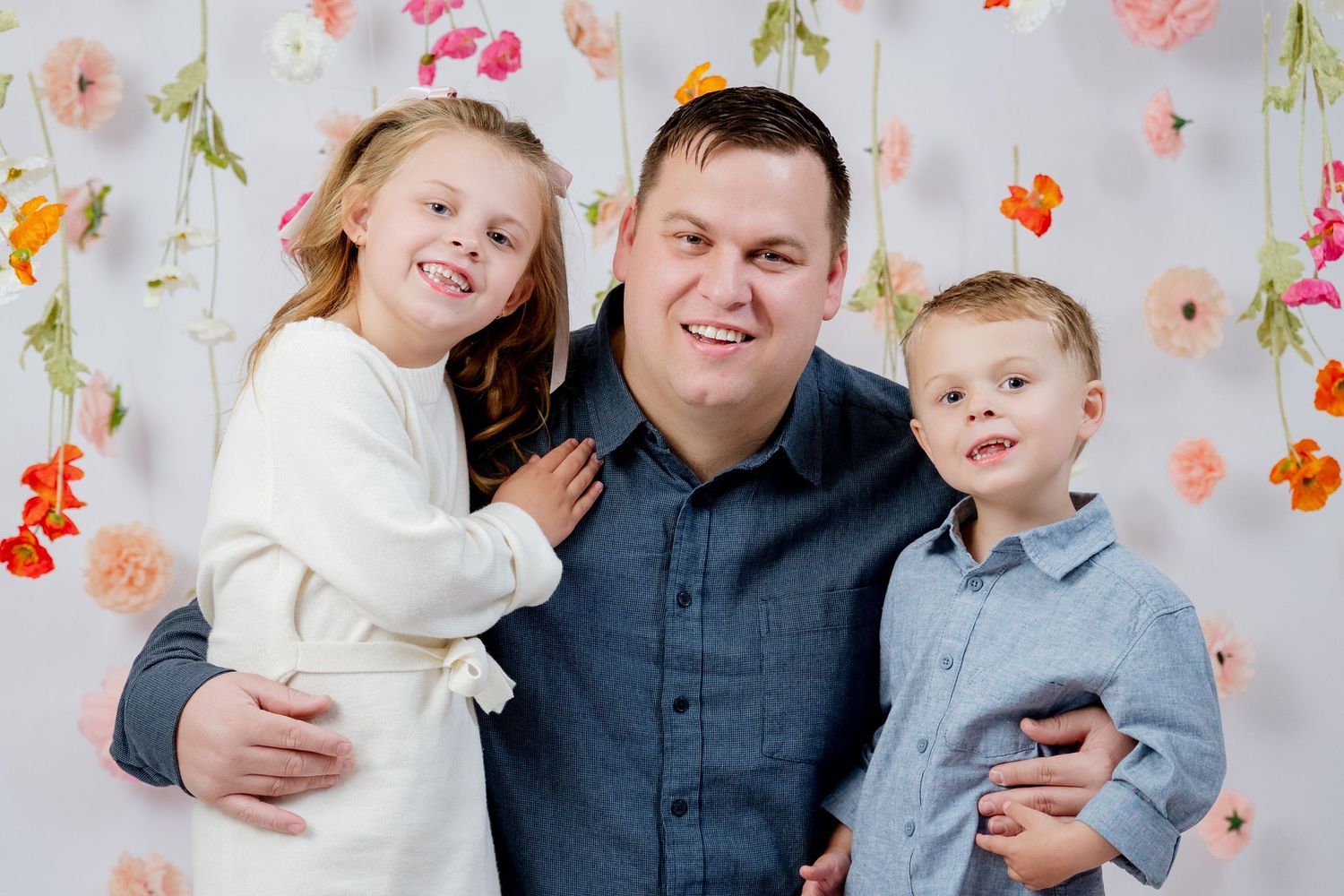 A man smiles with two young children in front of a floral backdrop in a professional family portrait.