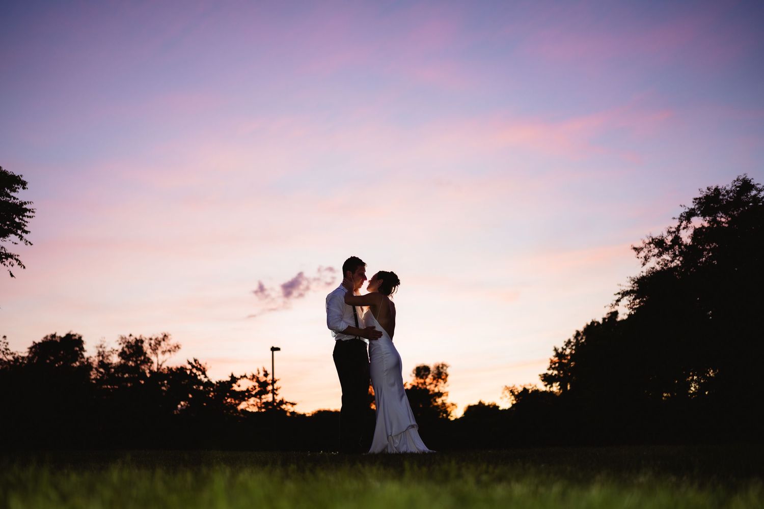 Silhouette of a bride and groom embracing at sunset with a colorful pink and purple sky backdrop.