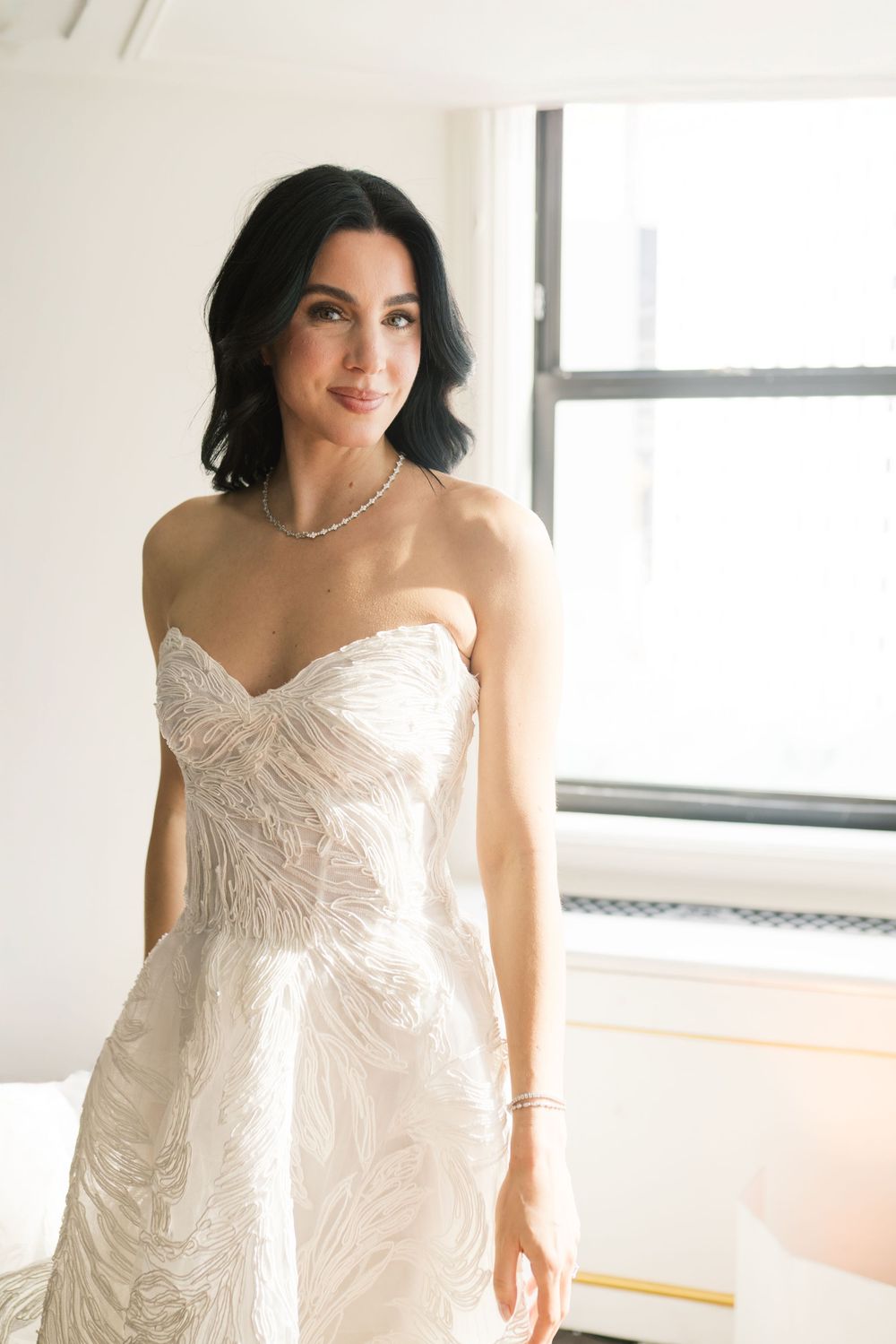 Bride in strapless embroidered white wedding gown with pearl necklace, posing elegantly near a bright window.