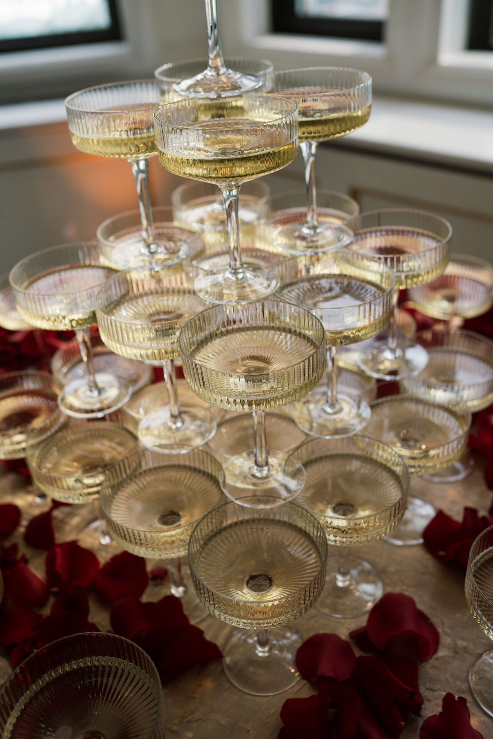 Elegant champagne tower with crystal coupe glasses filled with sparkling wine, surrounded by red rose petals at a wedding reception.