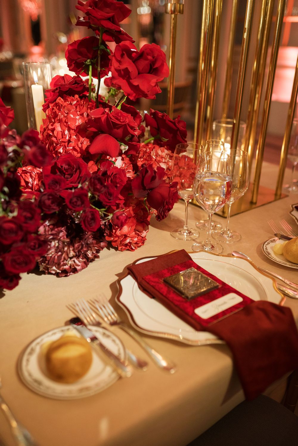 Elegant wedding table setting with red roses centerpiece, gold candles, crystal glasses, and crimson napkins on white linen.