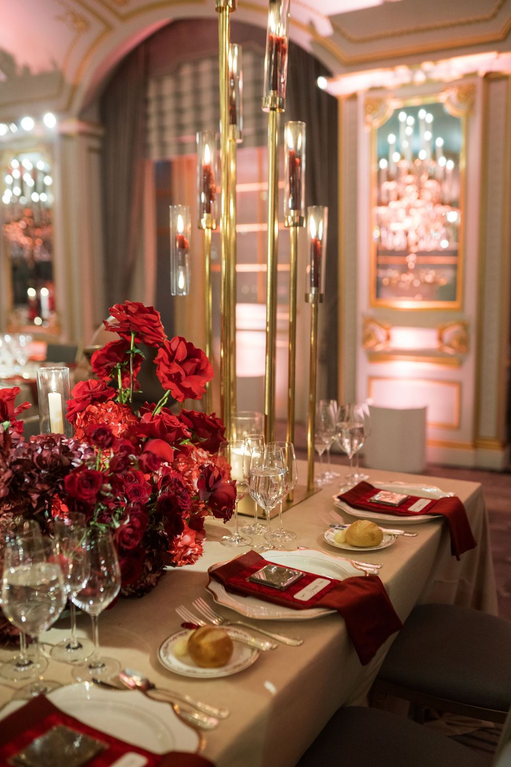 Elegant wedding reception table with red rose centerpieces, gold candelabras, and crimson napkins in a luxurious ballroom.