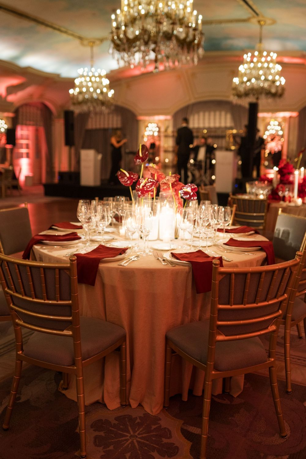 Elegant wedding reception with round tables set with red napkins, crystal glasses, floral centerpieces, and gold chairs under chandeliers.