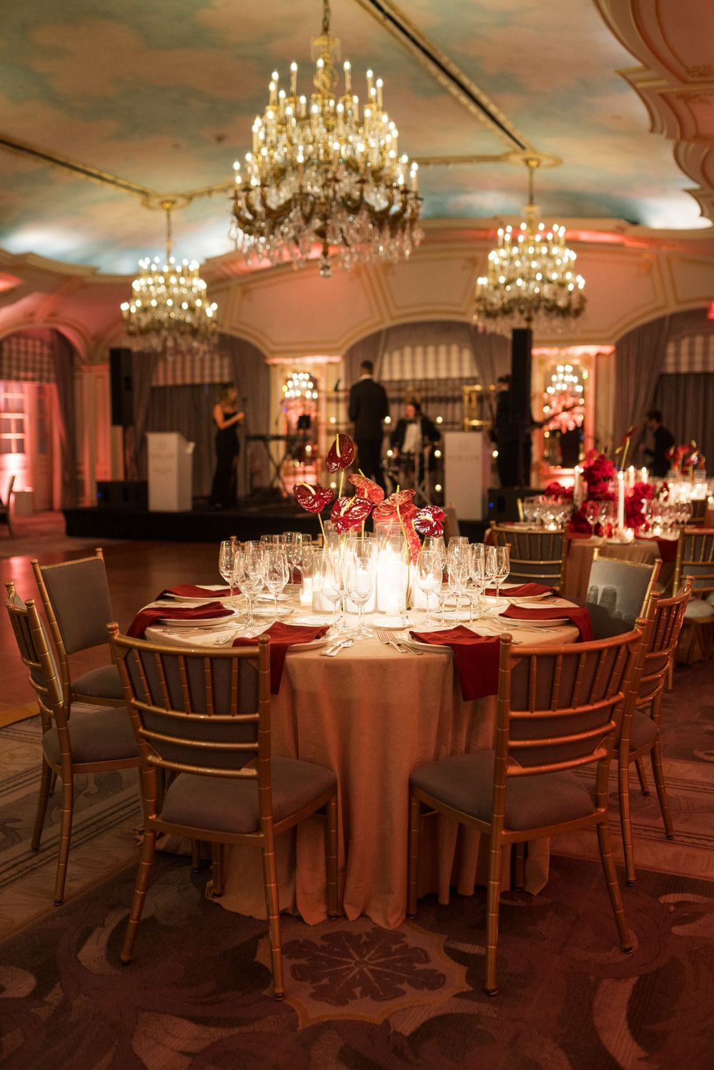 Elegant ballroom wedding reception with crystal chandeliers, gold chairs, red floral centerpieces, and candlelit round tables.