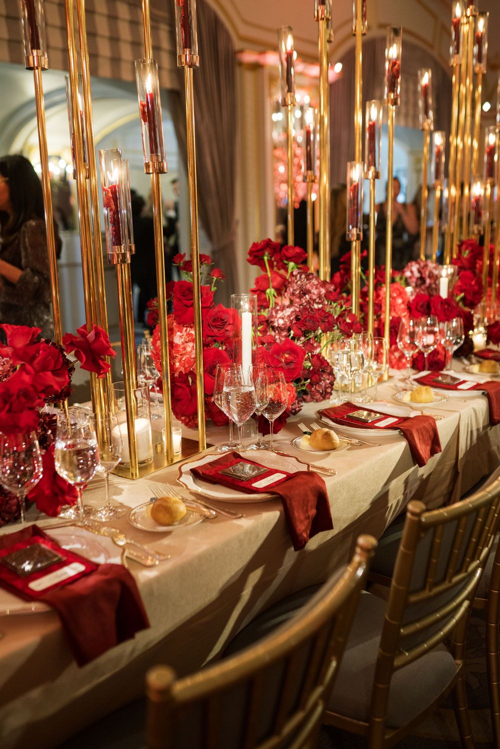 Elegant wedding reception table with red roses, gold candelabras, crimson napkins, and crystal glassware on a white linen tablecloth.