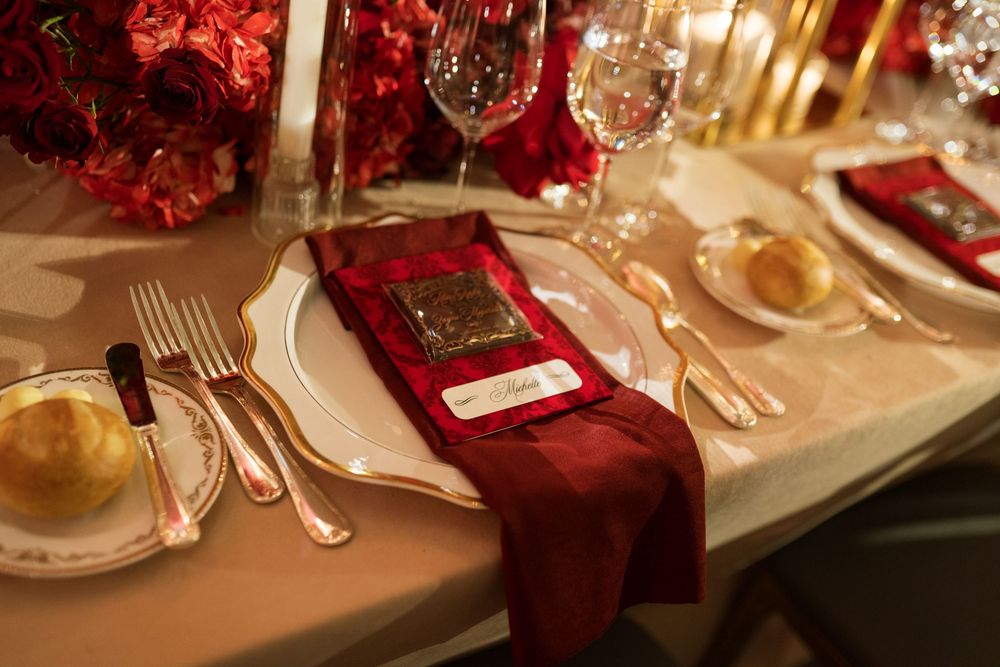 Elegant wedding table setting with crimson napkins, gold-rimmed plates, crystal glasses, and red floral centerpieces.