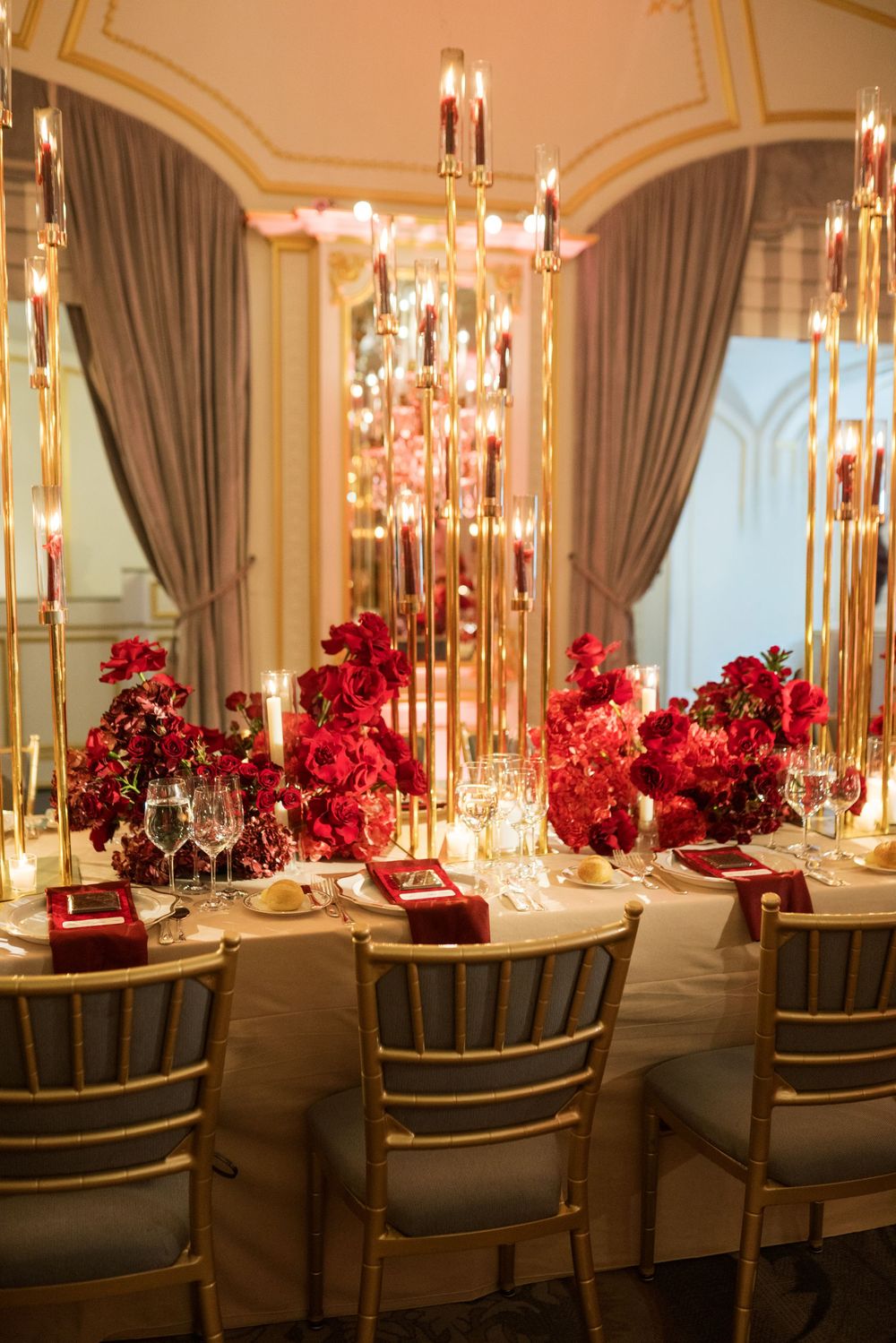Elegant wedding reception table with red rose centerpieces, gold candelabras, crystal glasses, and red napkins in a luxurious venue.