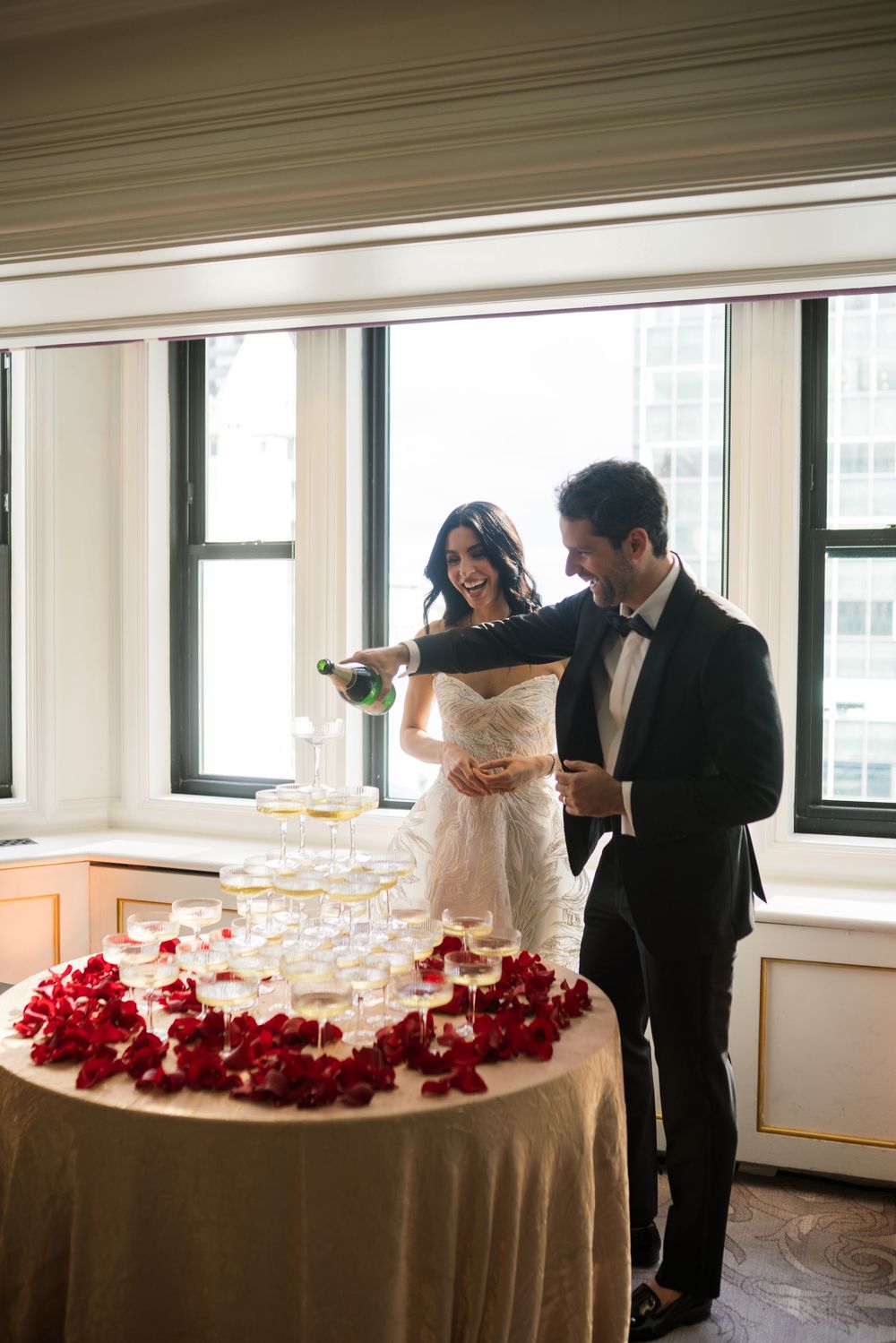 Bride and groom pouring champagne tower surrounded by red rose petals at elegant wedding reception.
