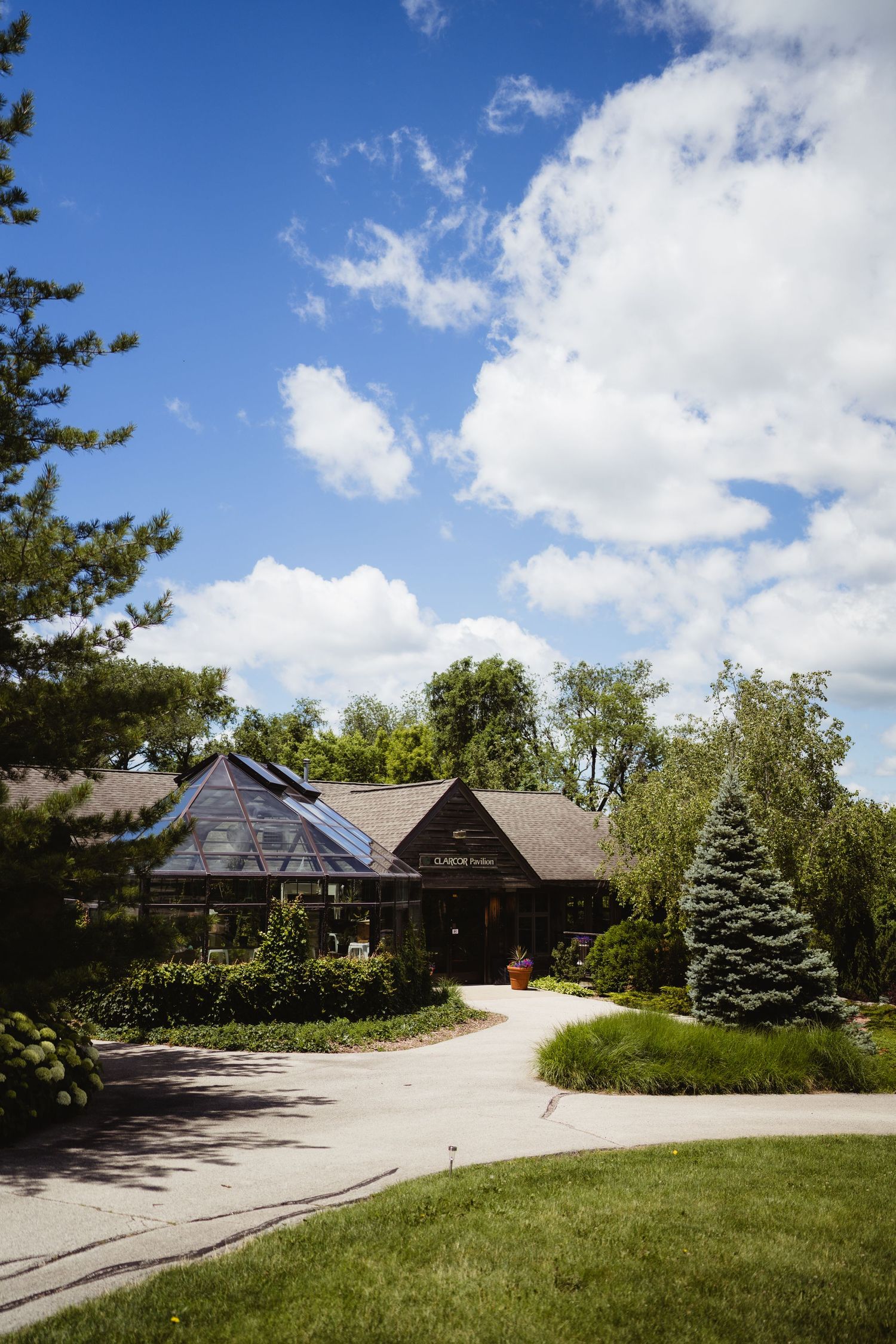 Charming greenhouse building surrounded by lush green trees and manicured lawns under a bright blue sky with fluffy clouds.