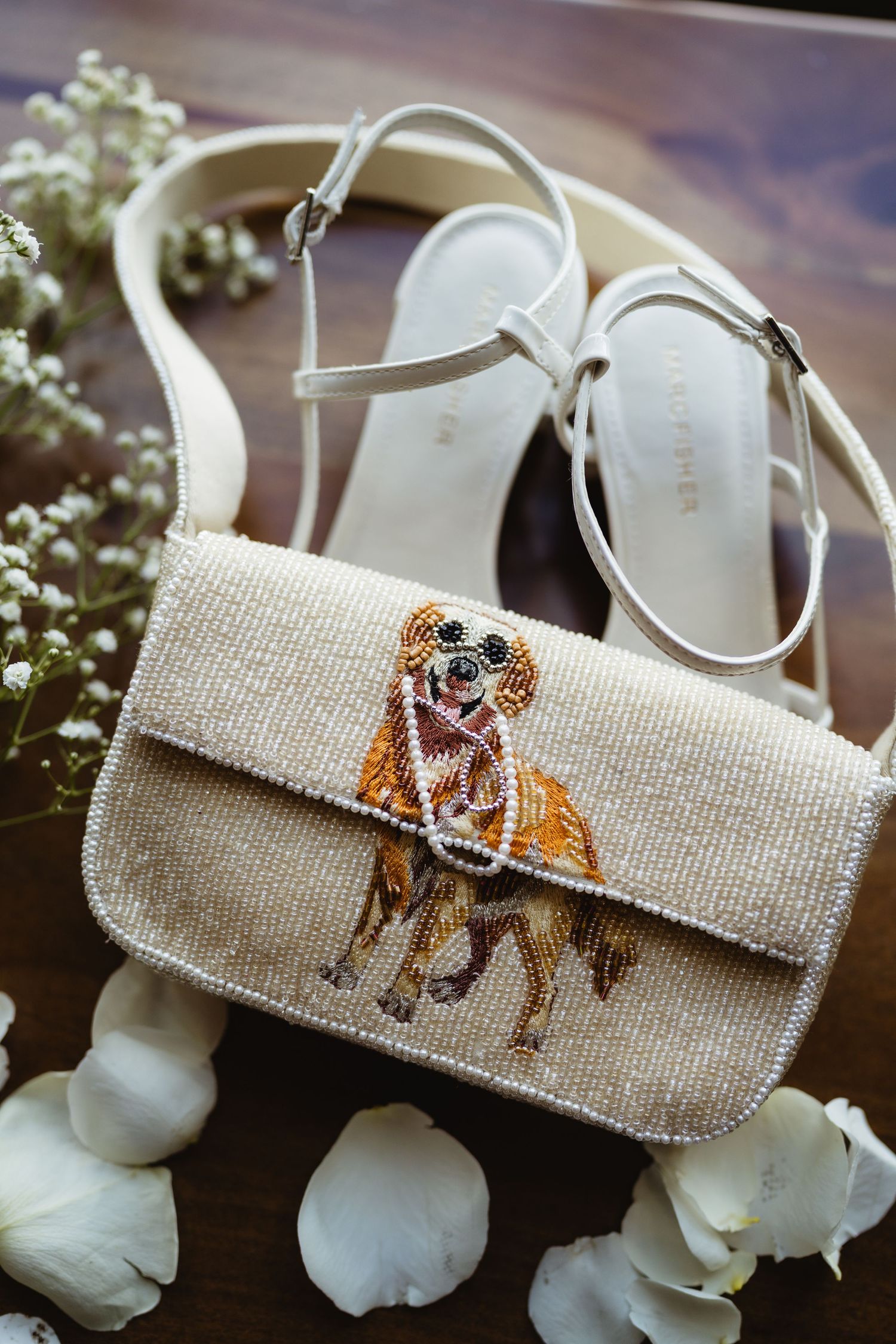 Beaded clutch purse with dog design beside white strappy heels and flower petals on wooden surface.