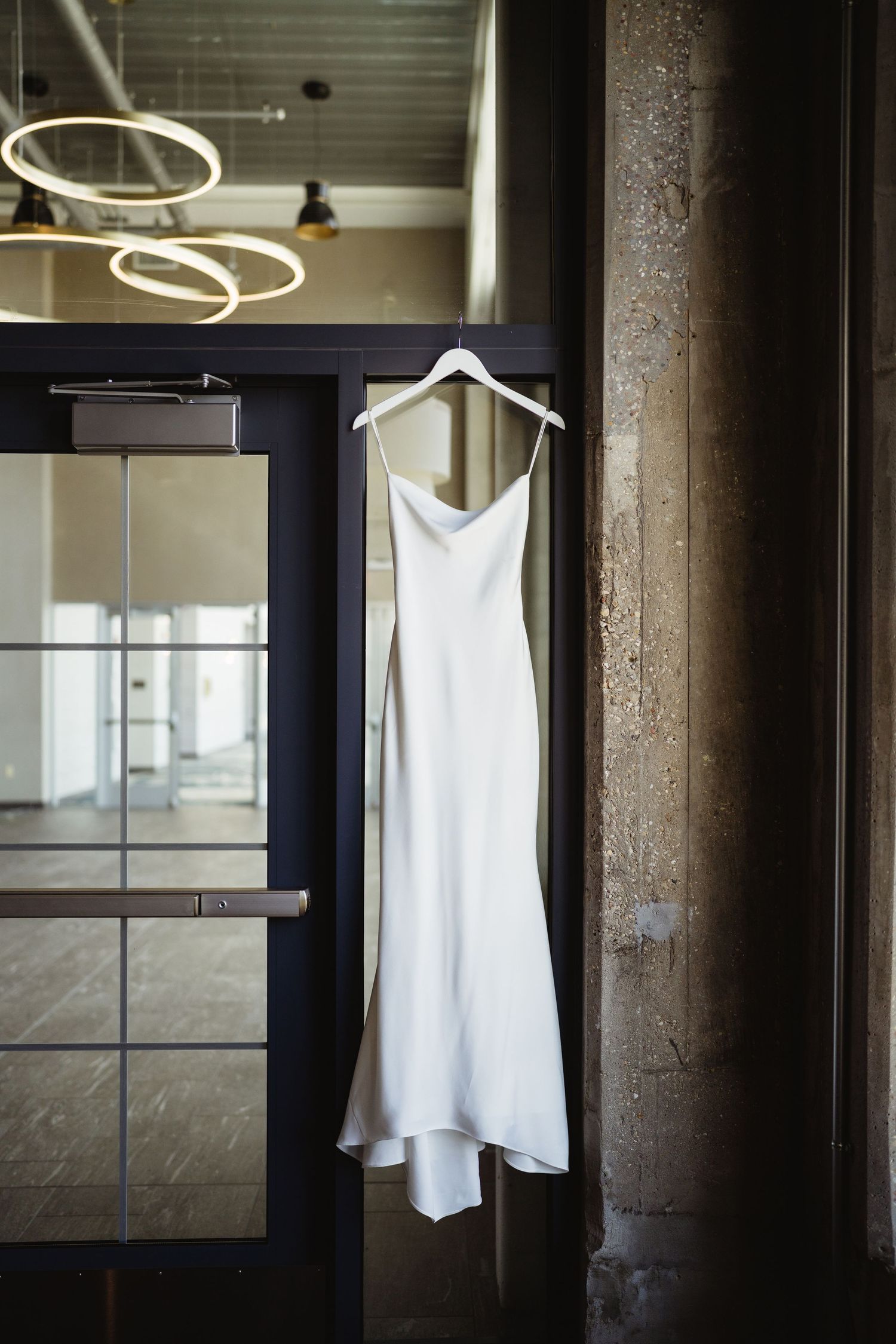 Elegant white satin slip wedding dress hanging on a hanger against a rustic column in a modern venue.