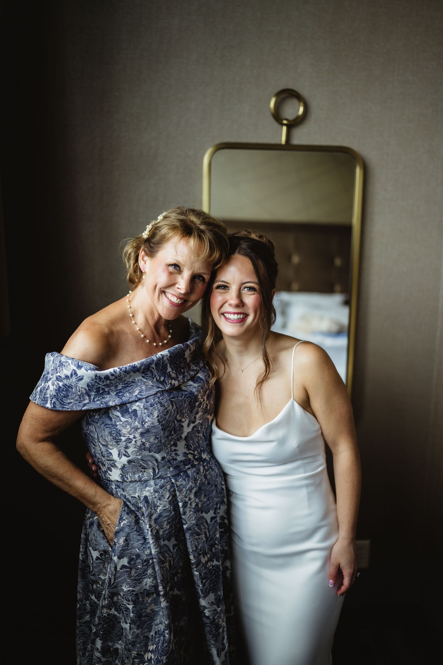 A bride in a white dress and older woman in blue floral gown smile together in front of a gold mirror.