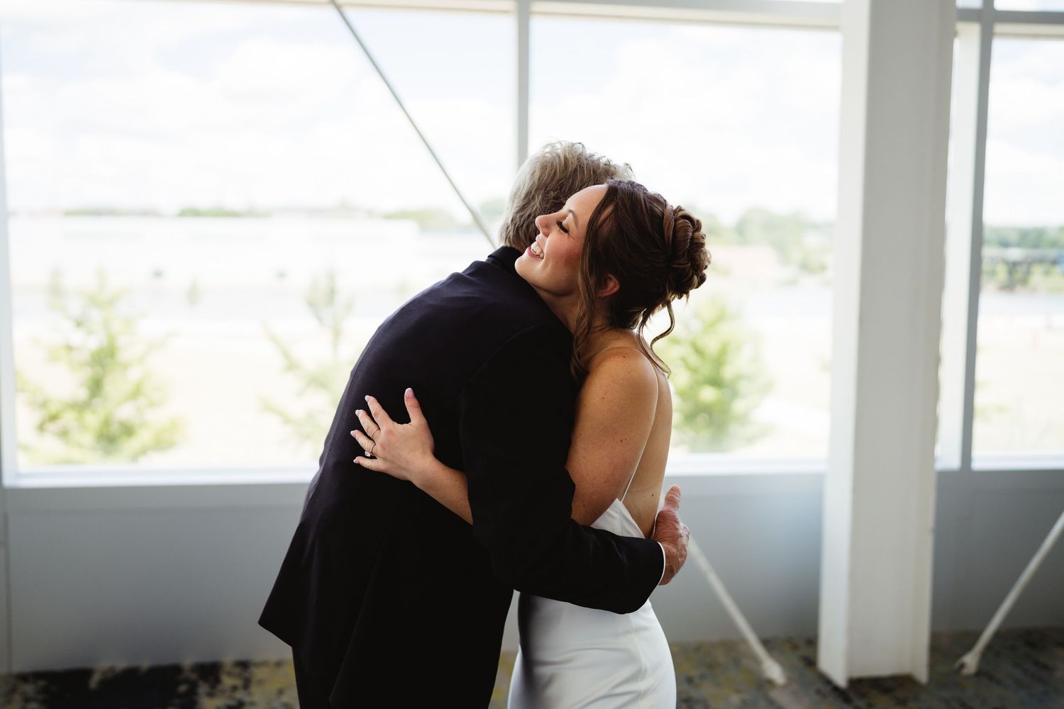 Bride in white gown shares an emotional hug with a guest in a dark suit near large windows at a wedding venue.