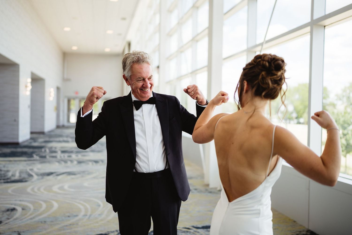 Father and bride share a joyful dancing moment in a bright venue, both flexing and celebrating on her wedding day.