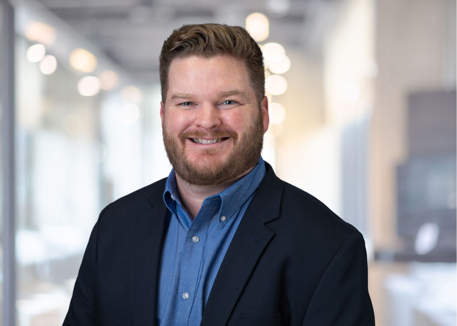 Smiling professional man with beard in blue shirt and blazer poses in modern office environment.
