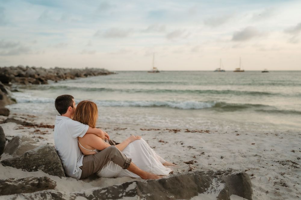 Romantic couple sitting together on rocky beach at sunset, watching sailboats on calm ocean waves.