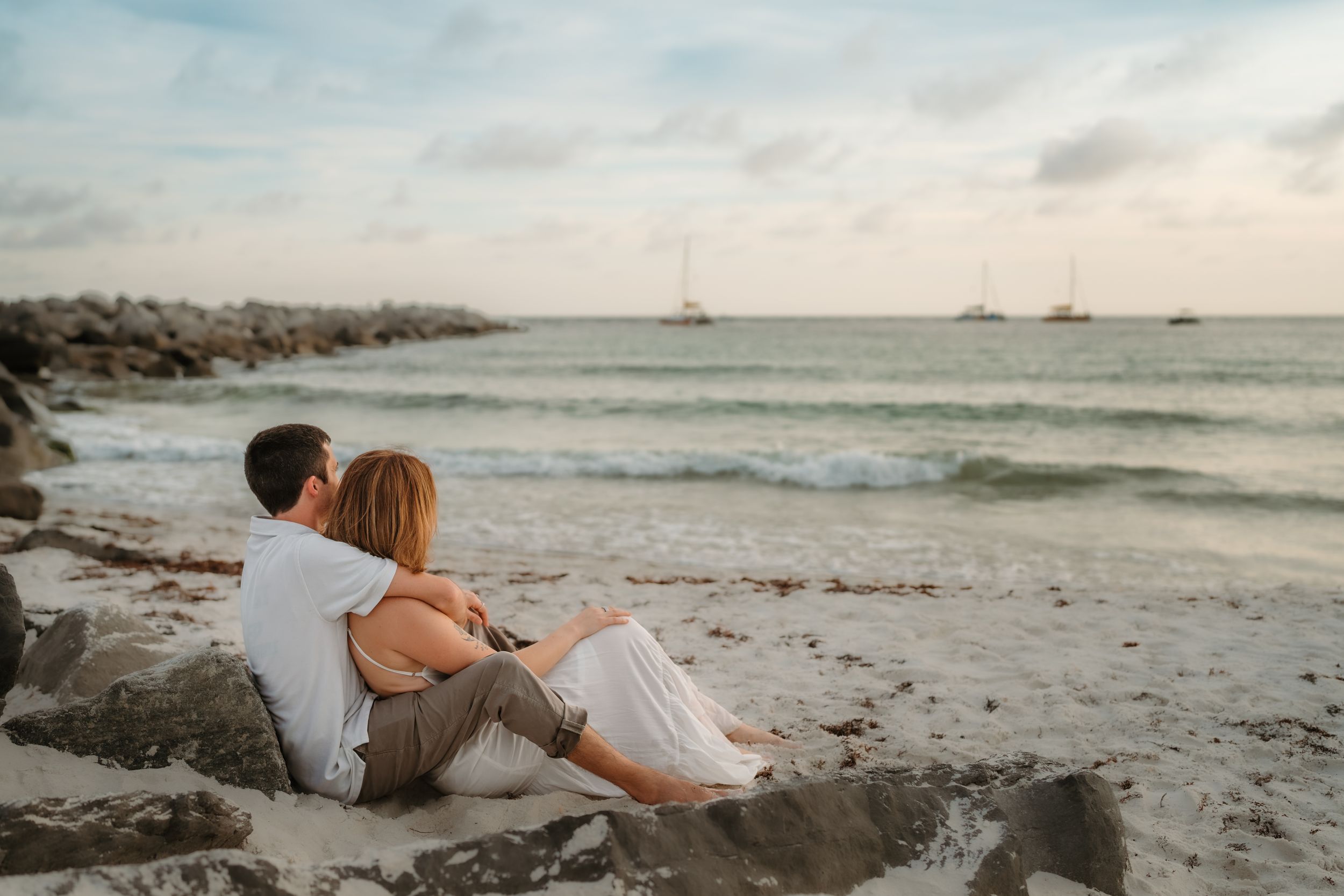 Romantic couple sitting together on rocky beach at sunset, watching sailboats on calm ocean waves.