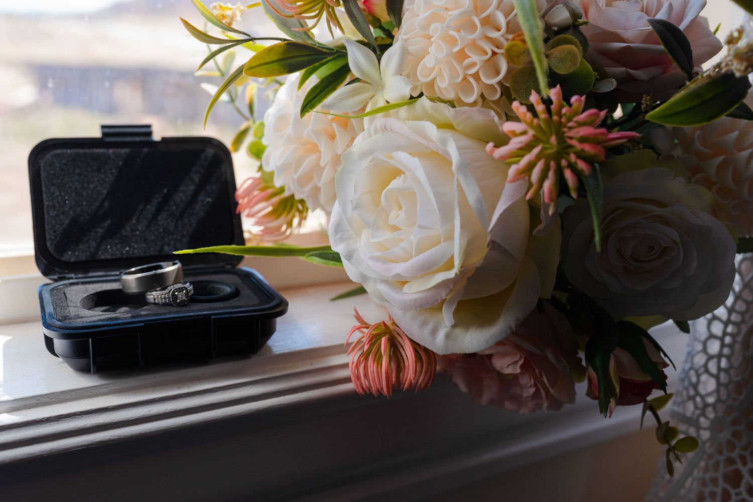 Wedding rings in a black velvet box beside a bridal bouquet of white roses and pink flowers on a windowsill.
