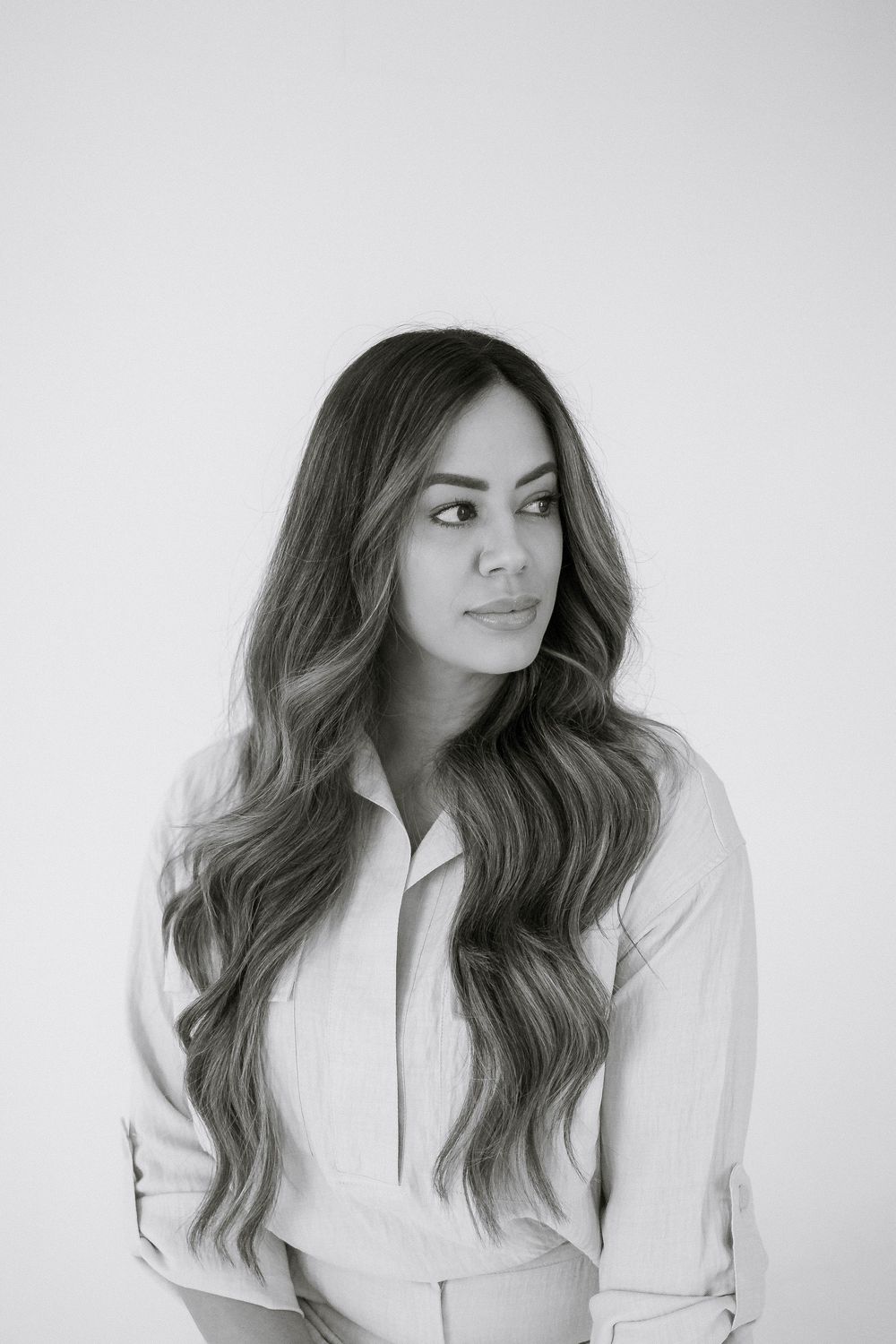 Black and white portrait of a woman with long wavy hair wearing a light button-up shirt, looking to the side.