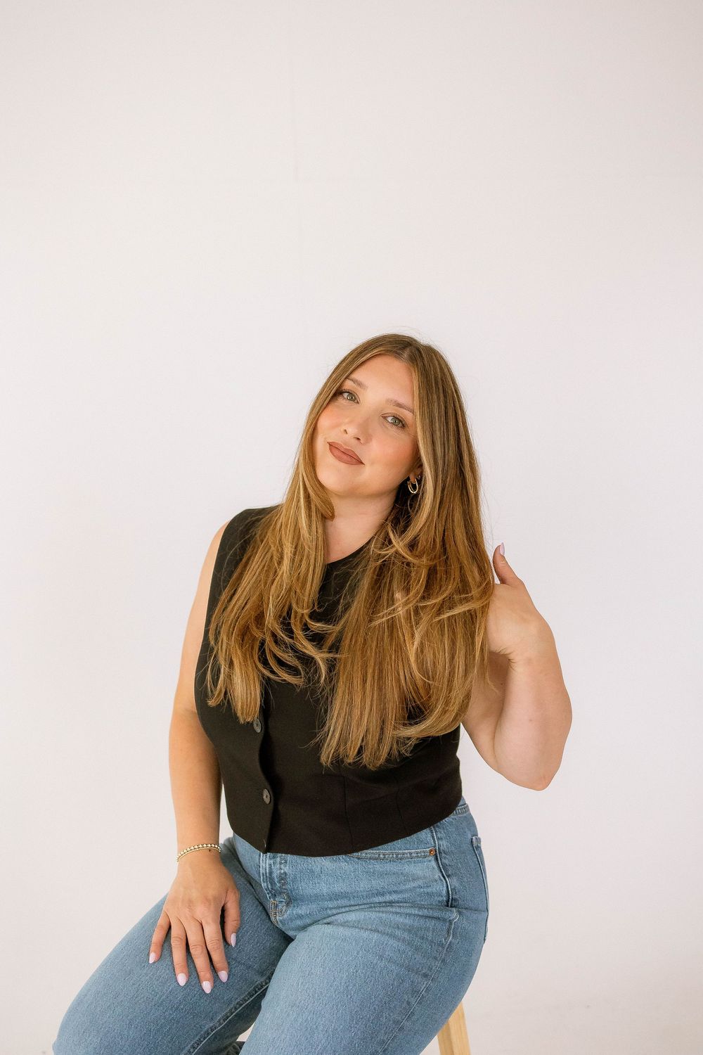 Young woman with long blonde hair sits confidently in black sleeveless top and blue jeans against white background.