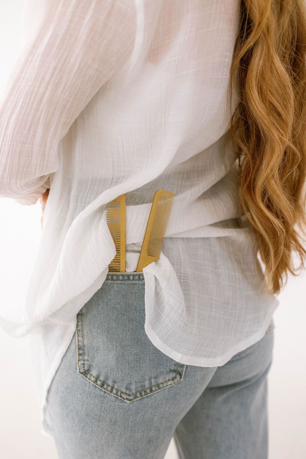 Close-up of gold hair scissors and comb tucked into back pocket of blue jeans with white linen shirt.