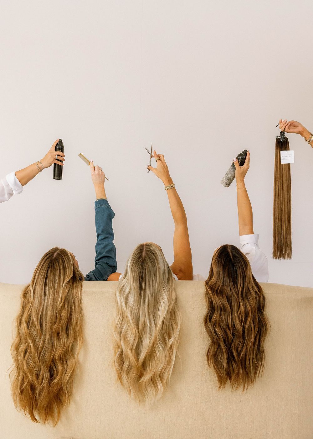 Three women with long wavy hair hold up hair tools and products, showcasing beautiful hair extensions and styling accessories.