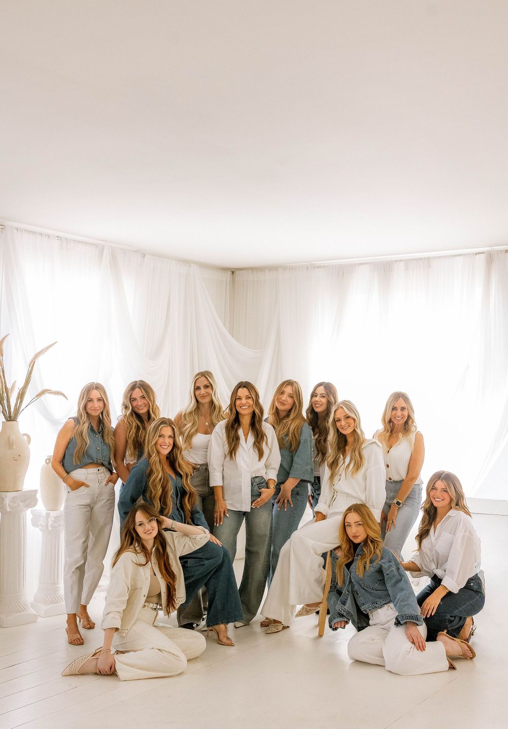 Group of women posing together in white and denim outfits in a bright, airy studio with white curtains.