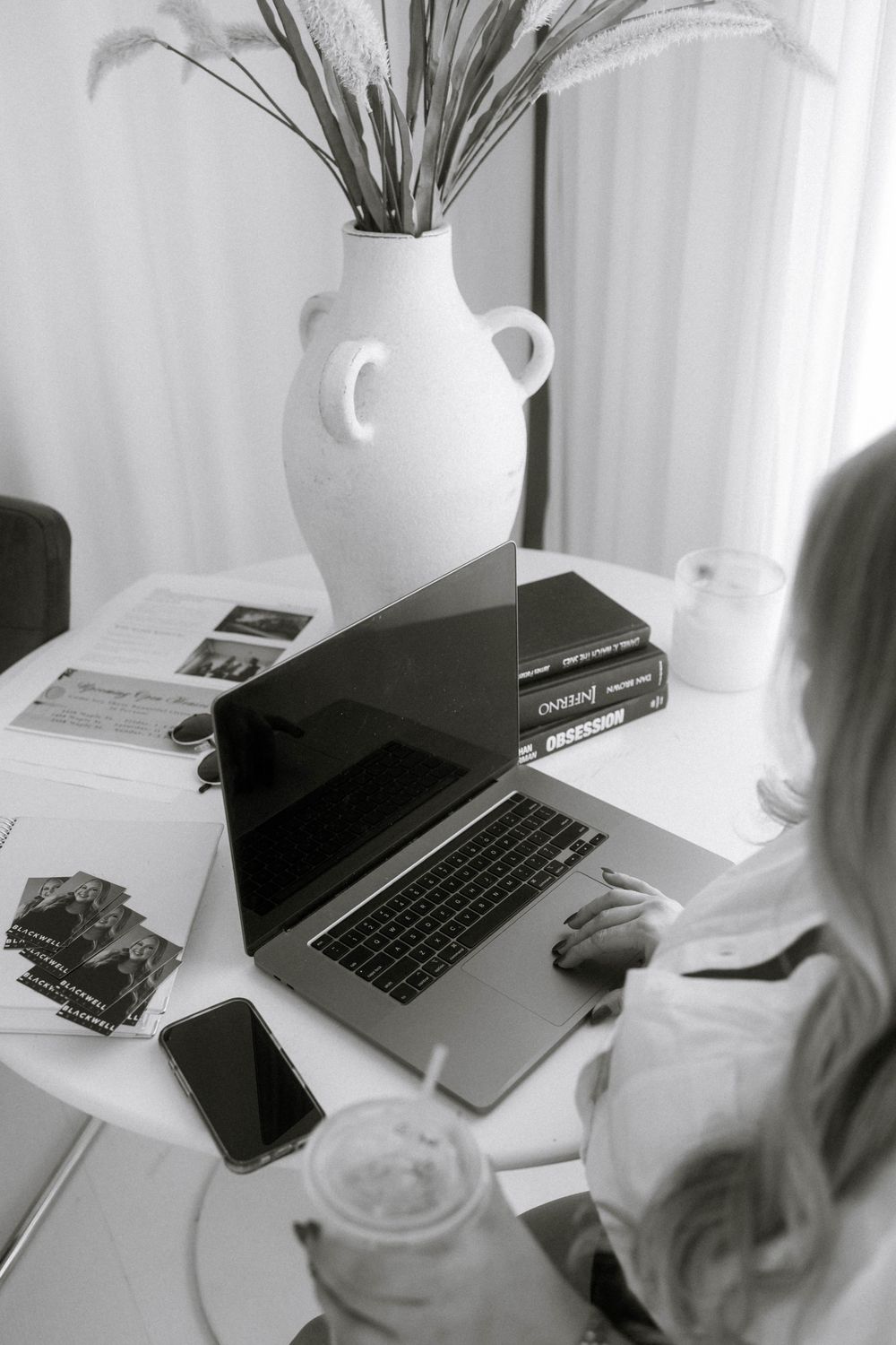 Black and white photo of a woman working on a laptop at a desk with a vase, books, phone, and iced coffee.