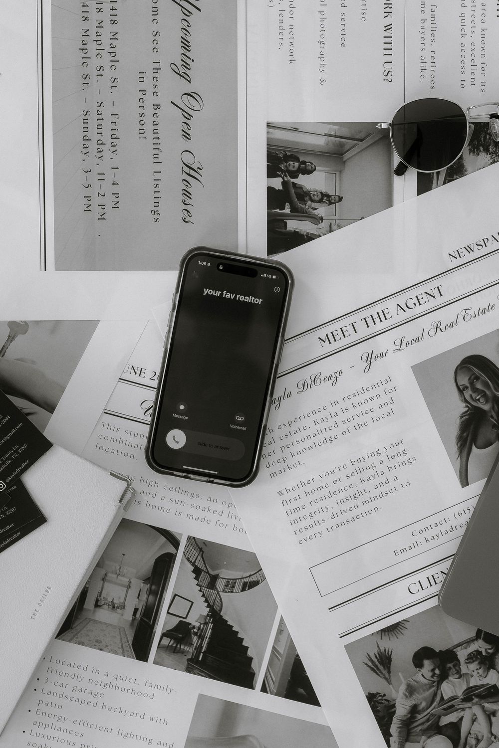 A smartphone displaying 'your fav realtor' rests on real estate marketing flyers and a newspaper beside sunglasses.