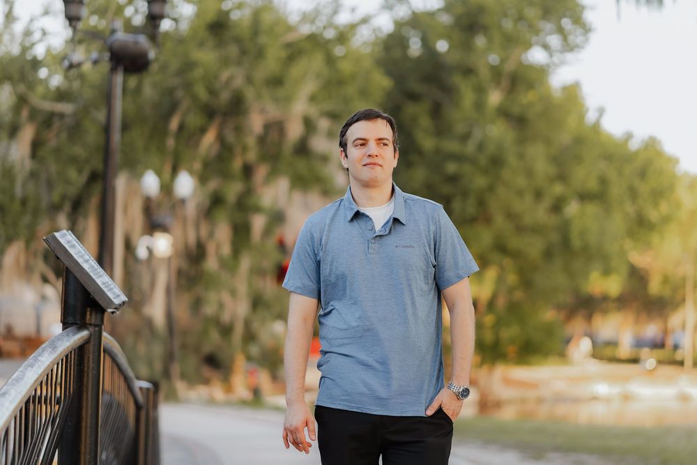 Man in blue polo shirt walking outdoors on a tree-lined path in a sunny park setting.