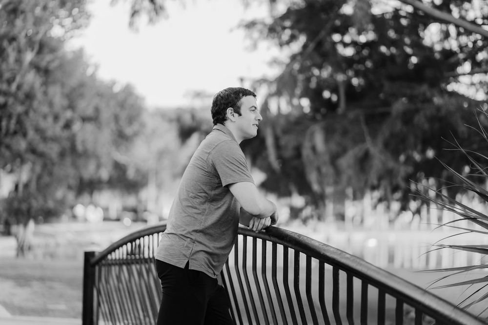 A young man leans on a railing of a bridge in a park, gazing into the distance in a black and white photo.