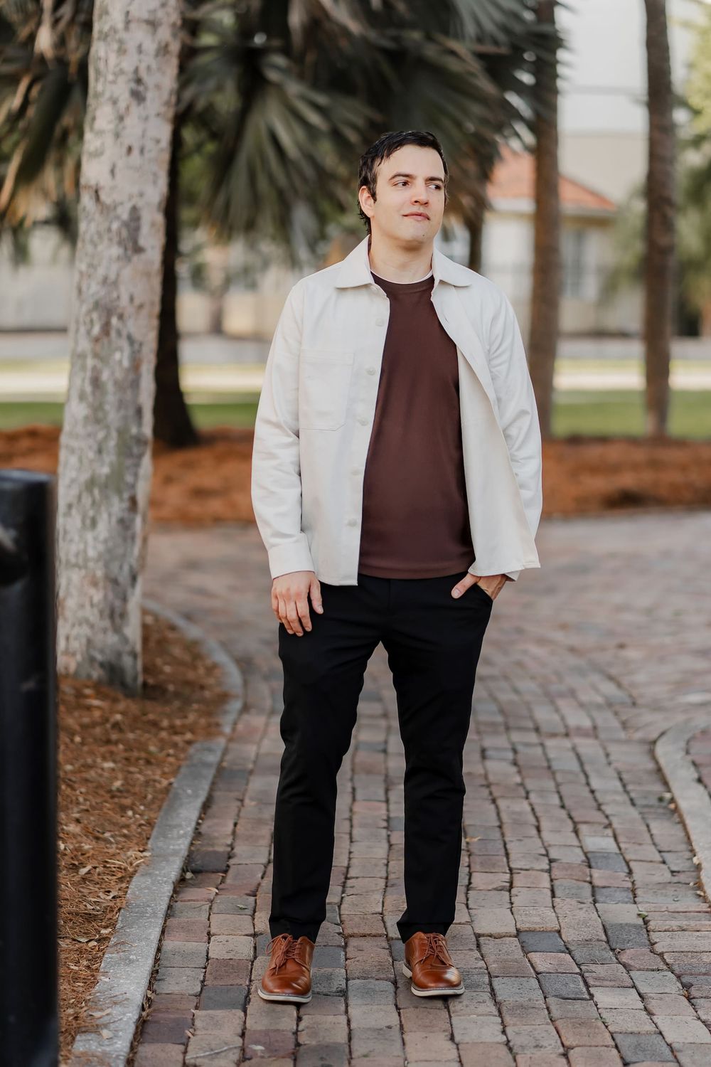 Young man in cream jacket and burgundy shirt standing on brick pathway surrounded by palm trees.