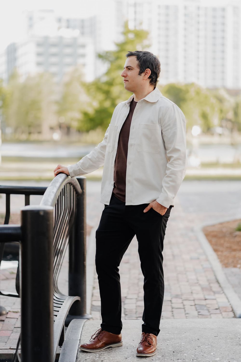 A well-dressed man in white shirt and black pants leans on a bench outdoors in an urban setting.