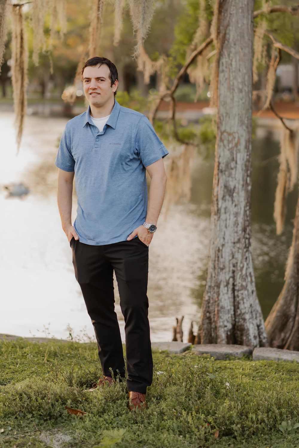 Young man in blue polo and black pants standing by a serene lake with Spanish moss-draped trees.