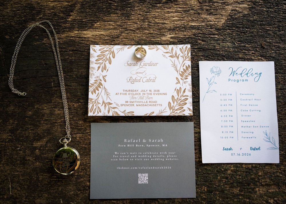 Wedding stationery flat lay featuring gold leaf invitation, program, dark envelope, and vintage pocket watch on wood.