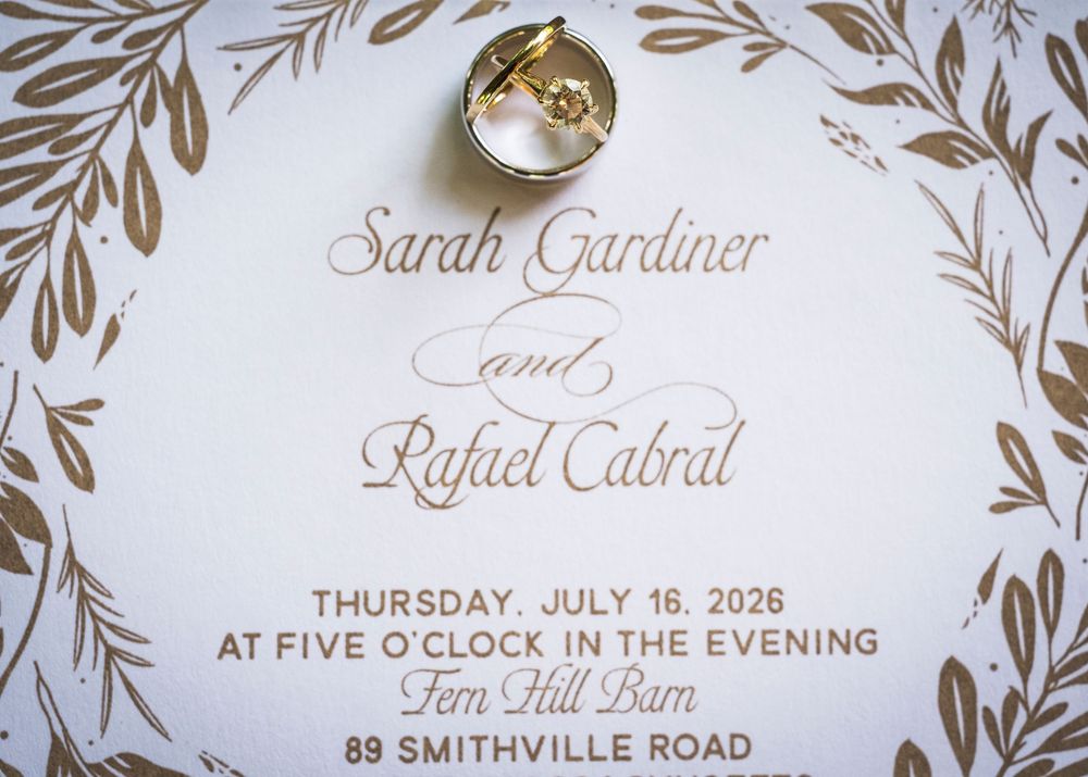 Wedding invitation for Sarah Gardiner and Rafael Cabral at Fern Hill Barn with gold rings resting on top.