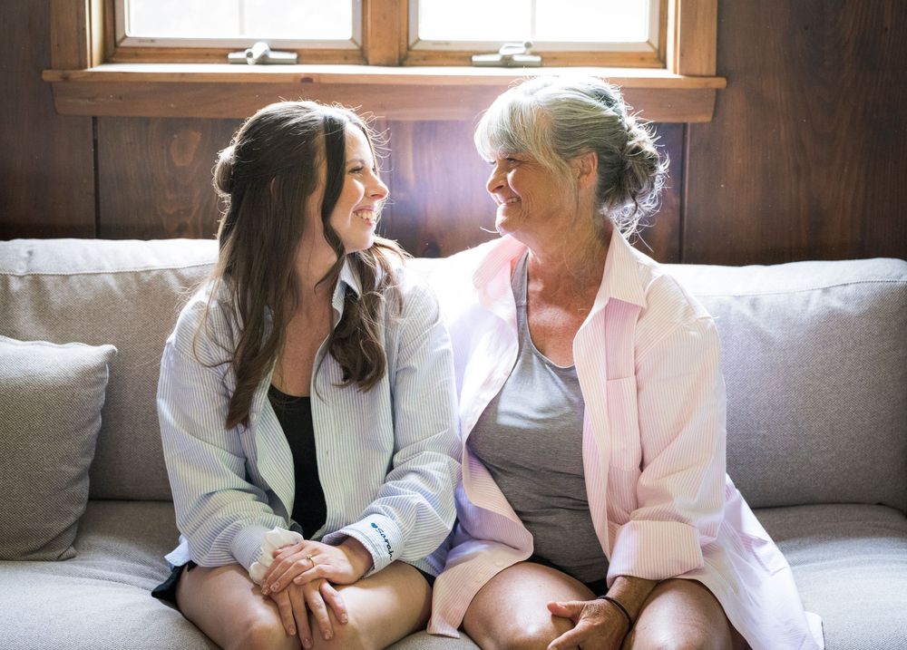 Two women share a warm smile while sitting together on a cozy couch in a sunlit room.