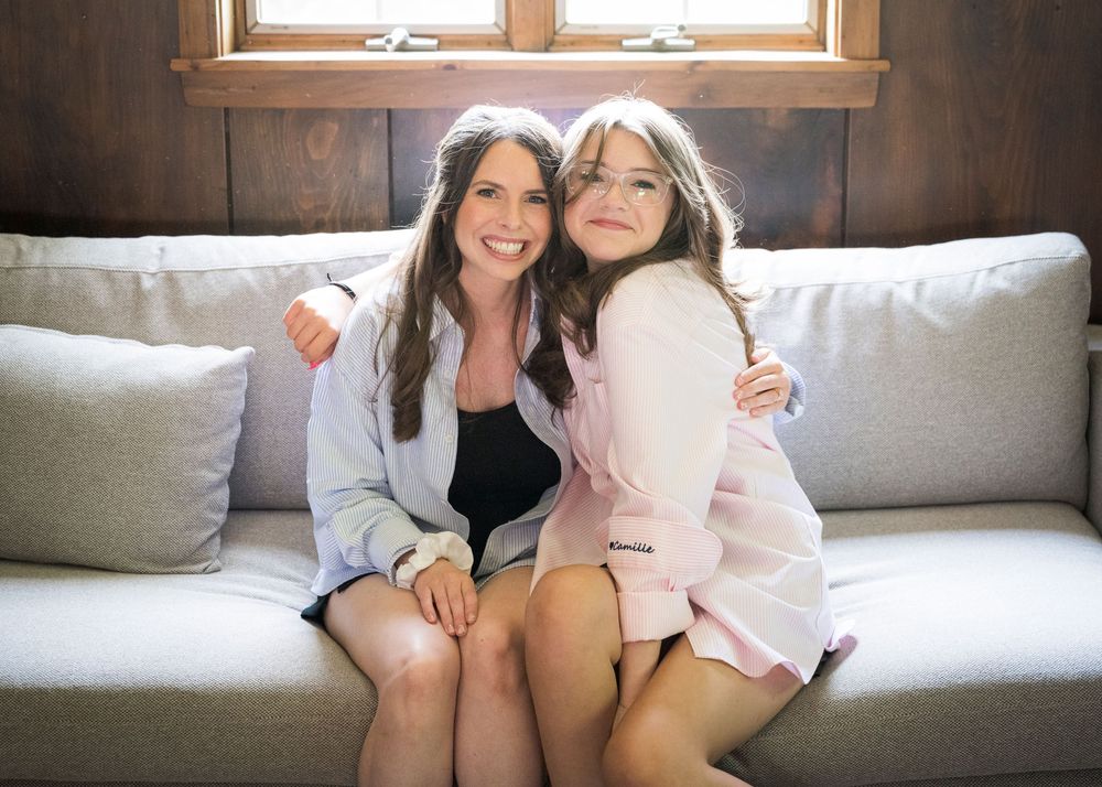 Two smiling bridesmaids in white and pink blazers sitting together on a gray couch in a wooden cabin.
