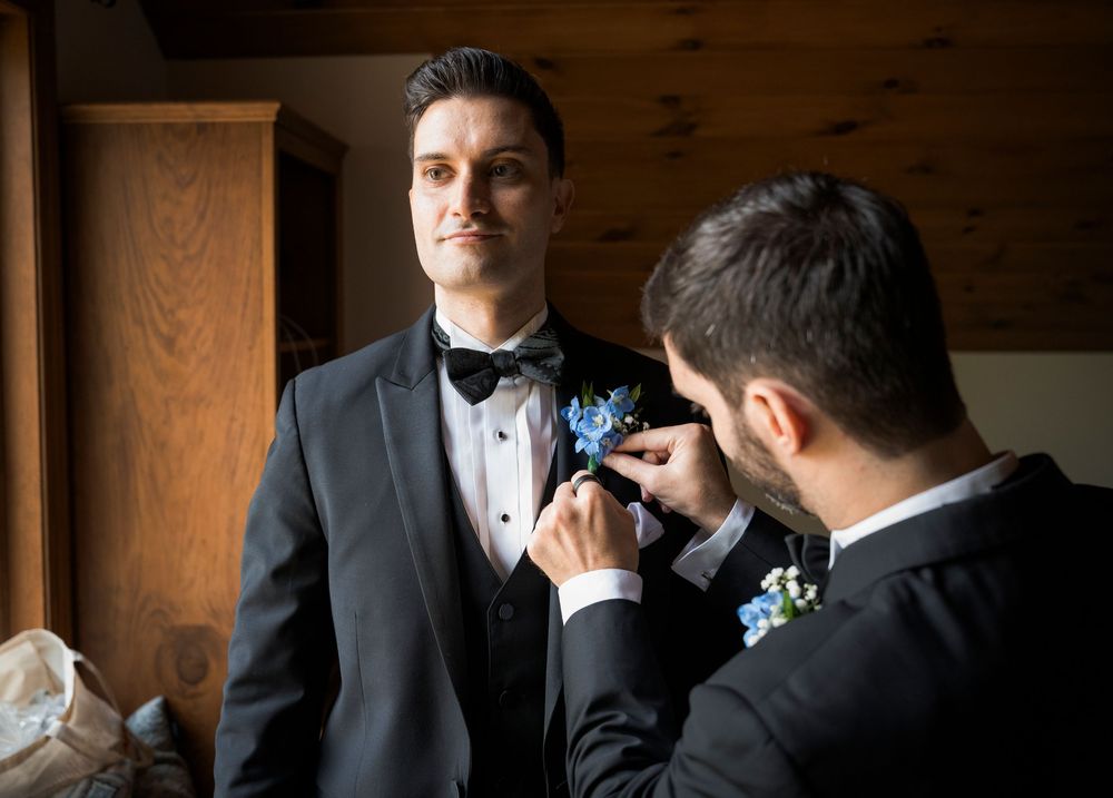 Groomsman pinning blue boutonniere on groom in black tuxedo before wedding ceremony.