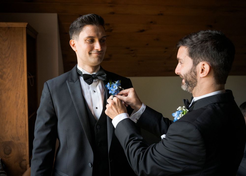 Two groomsmen in black tuxedos pin boutonnieres before a wedding ceremony.