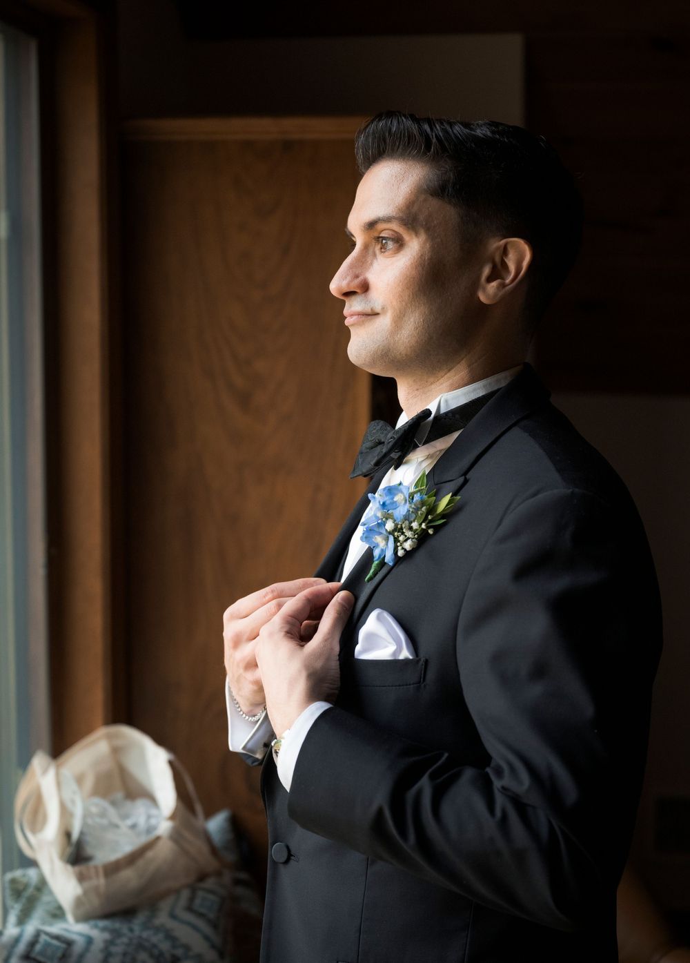 Groom in black tuxedo adjusting bow tie near window with blue floral boutonniere.