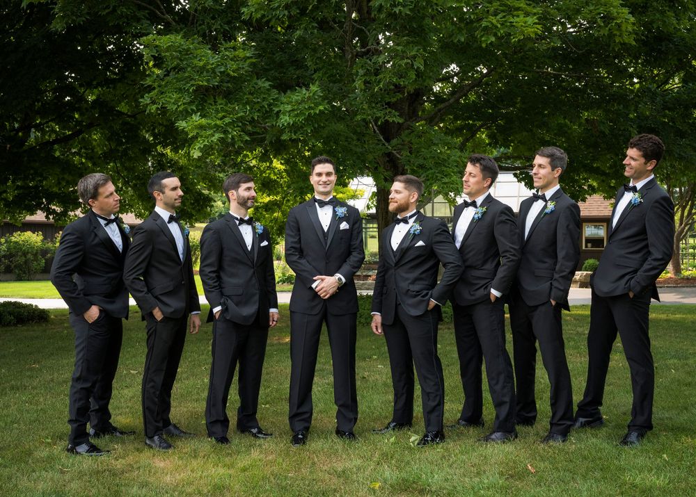 Groom and seven groomsmen in matching black tuxedos standing on green lawn outdoors.