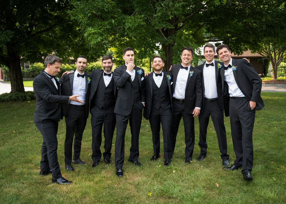 Eight groomsmen in black tuxedos with bow ties pose together outdoors on green grass at a wedding.