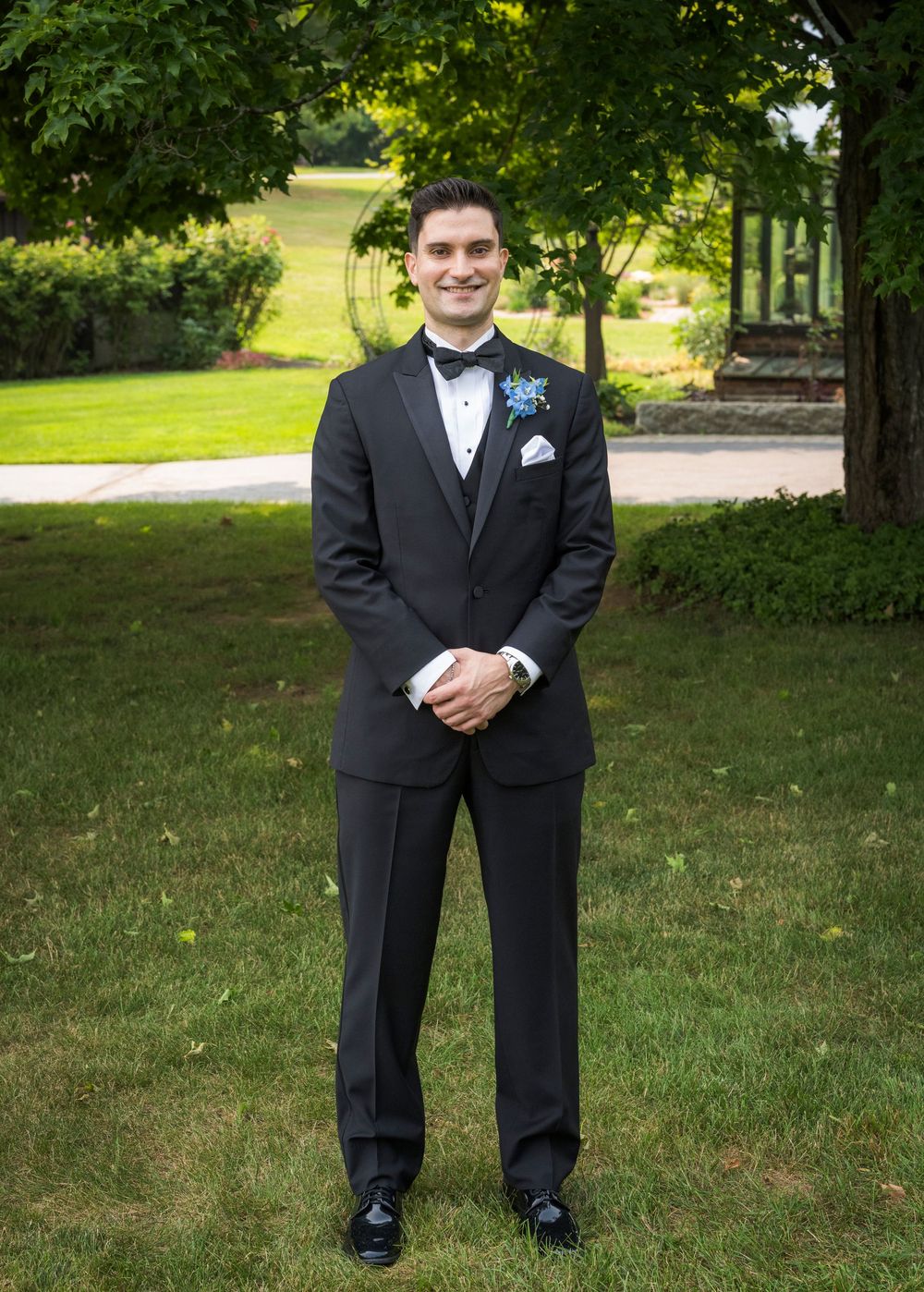 Groom in black tuxedo with blue boutonniere standing on green lawn at outdoor wedding venue.