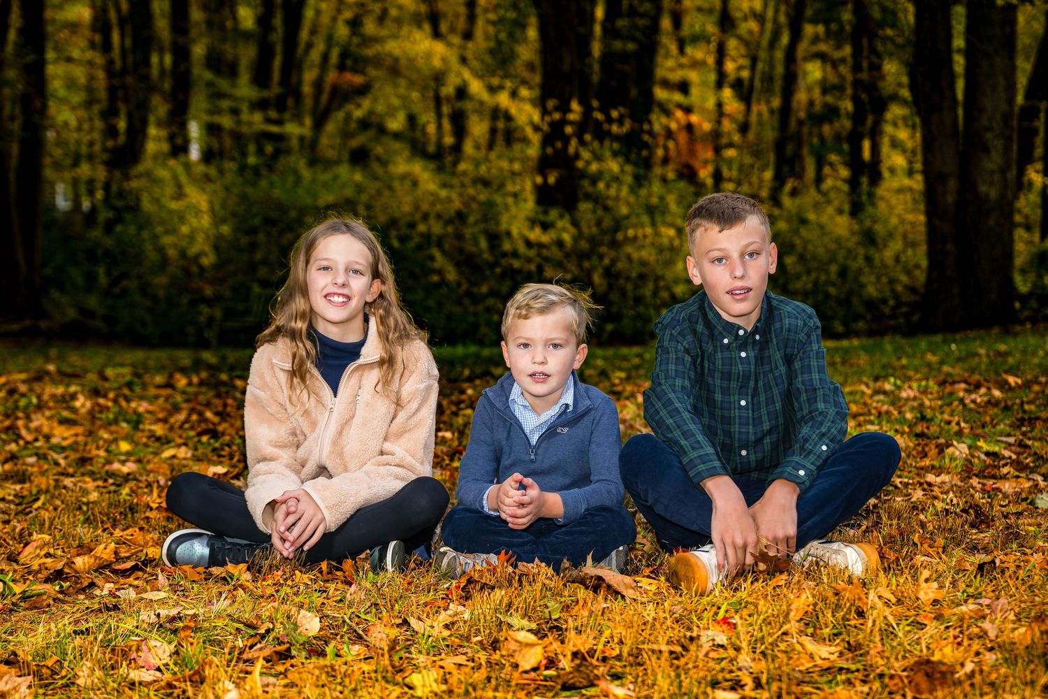 Three children sitting on autumn leaves in a park surrounded by colorful fall foliage trees.
