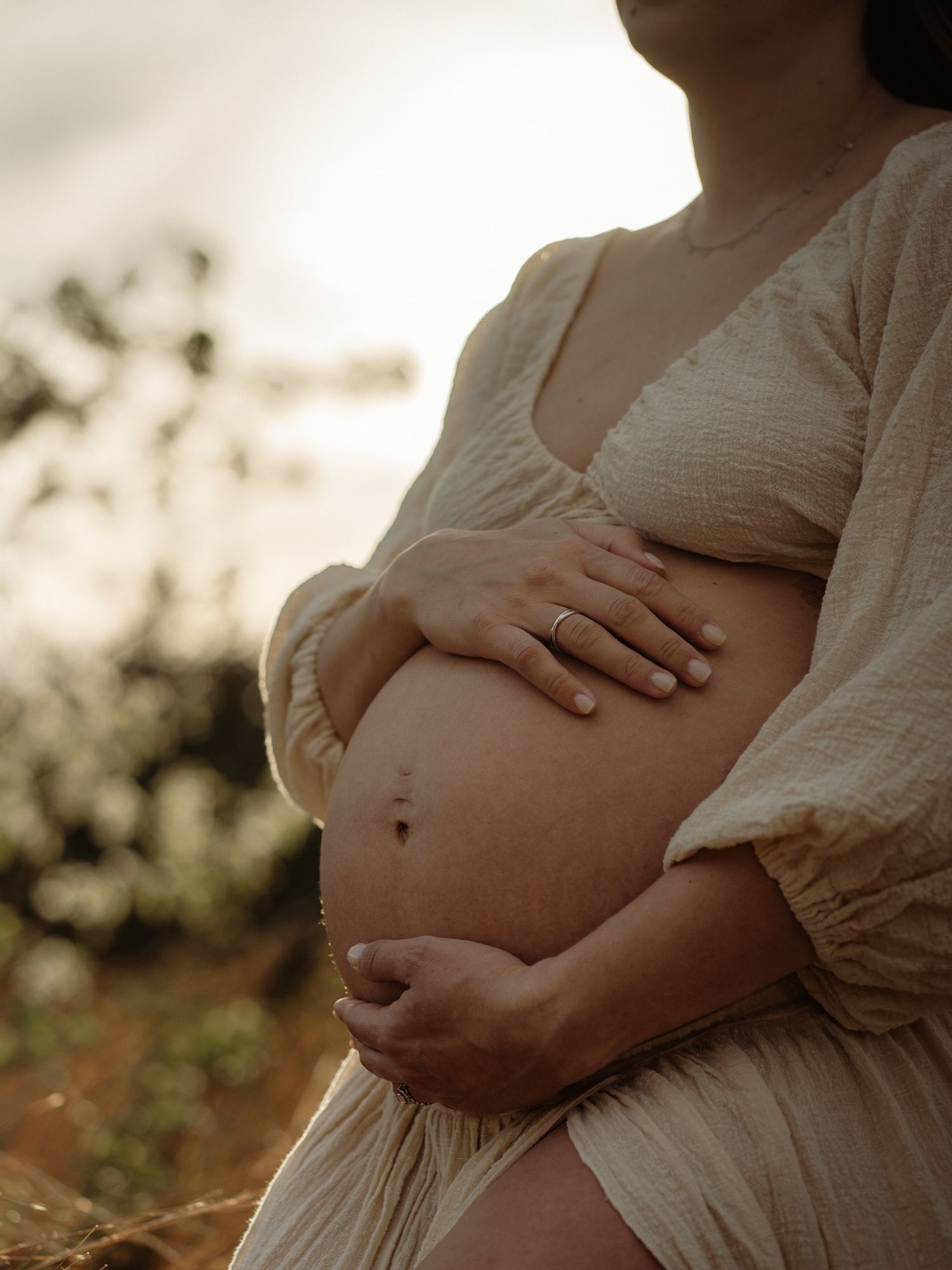 Pregnant woman in sheer white dress holding wildflowers in golden hour sunlight among blooming trees.