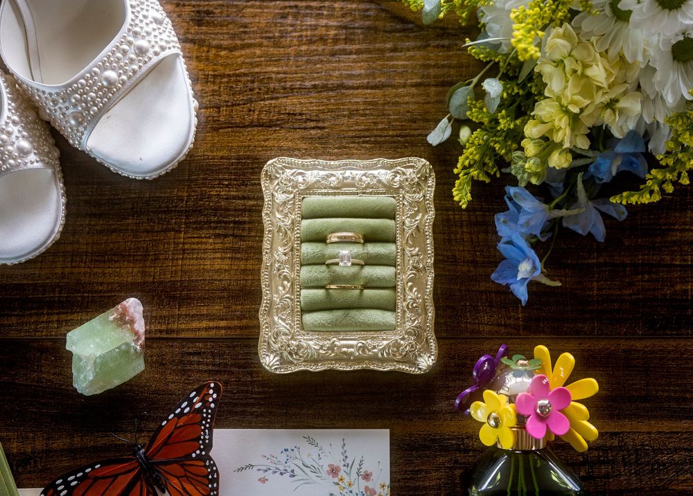 Wedding rings displayed on green velvet in ornate gold frame, with bridal heels, butterfly card, and colorful flowers on wood.