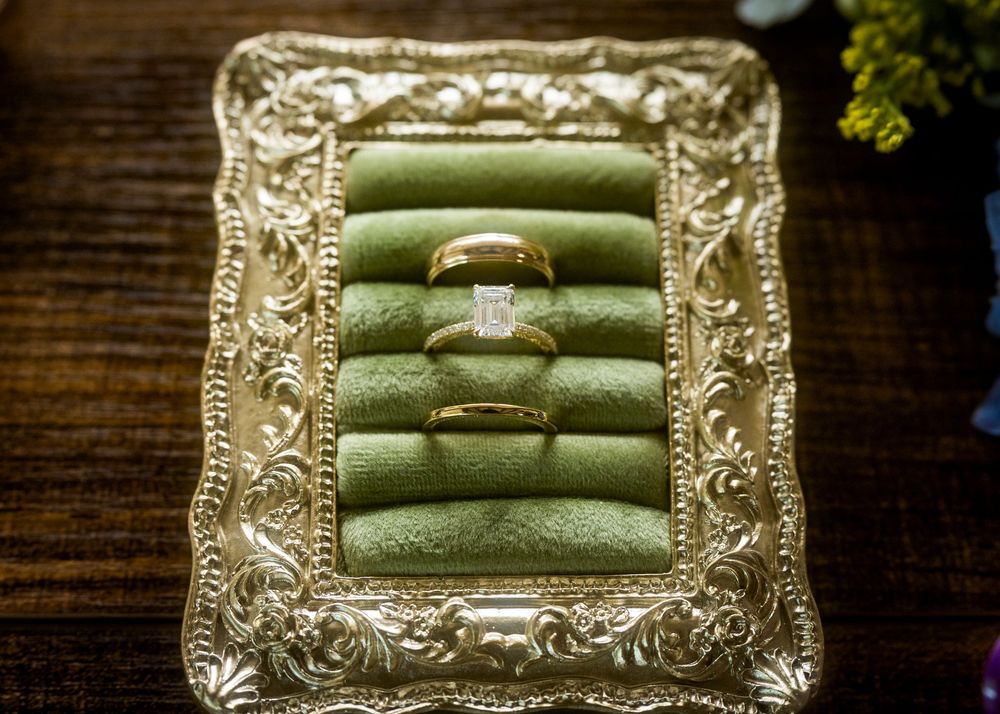 Gold wedding rings displayed on a green velvet tray in an ornate gold frame on a wooden surface.