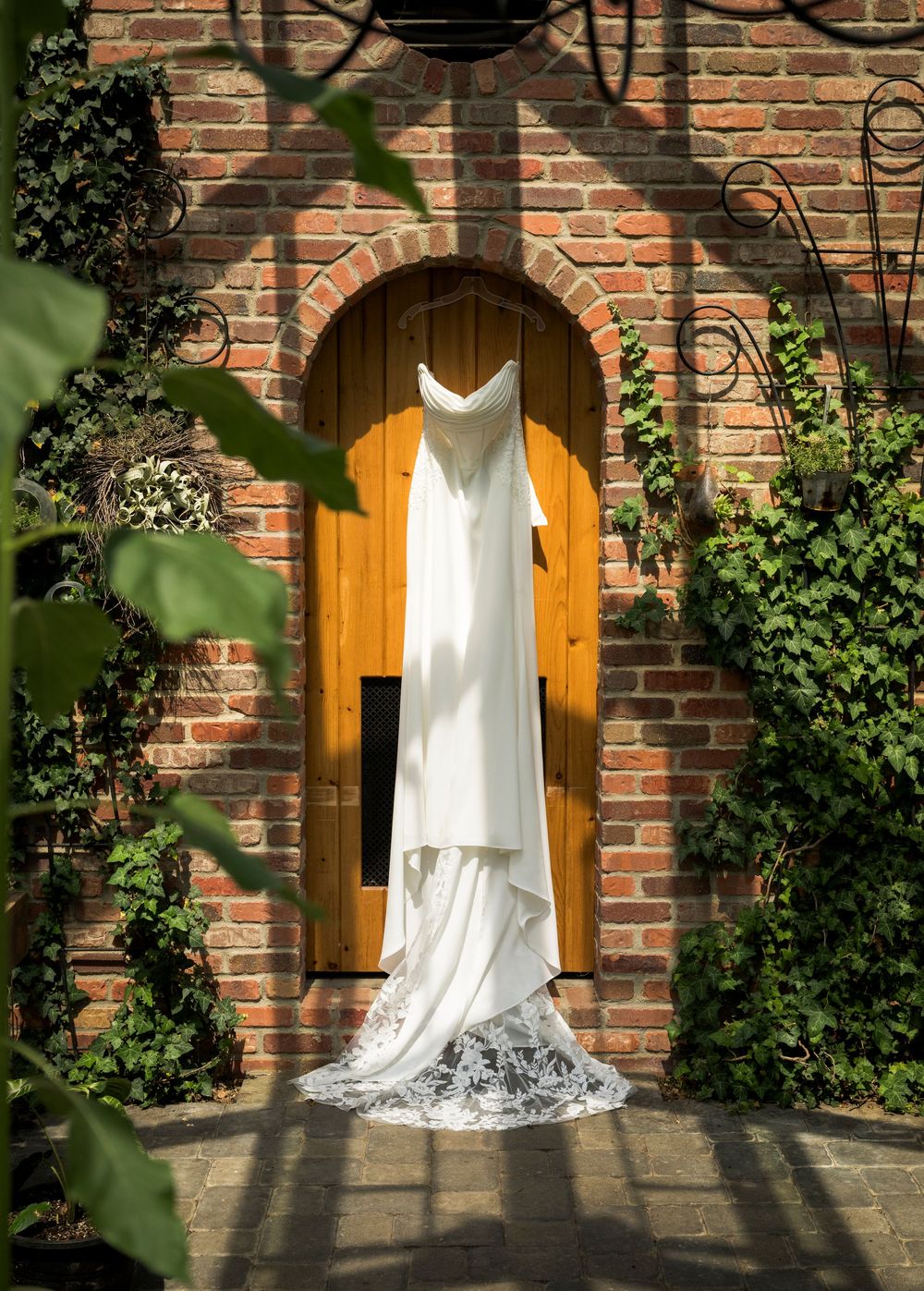 Elegant white lace wedding gown hanging in a brick archway with a wooden door and ivy.