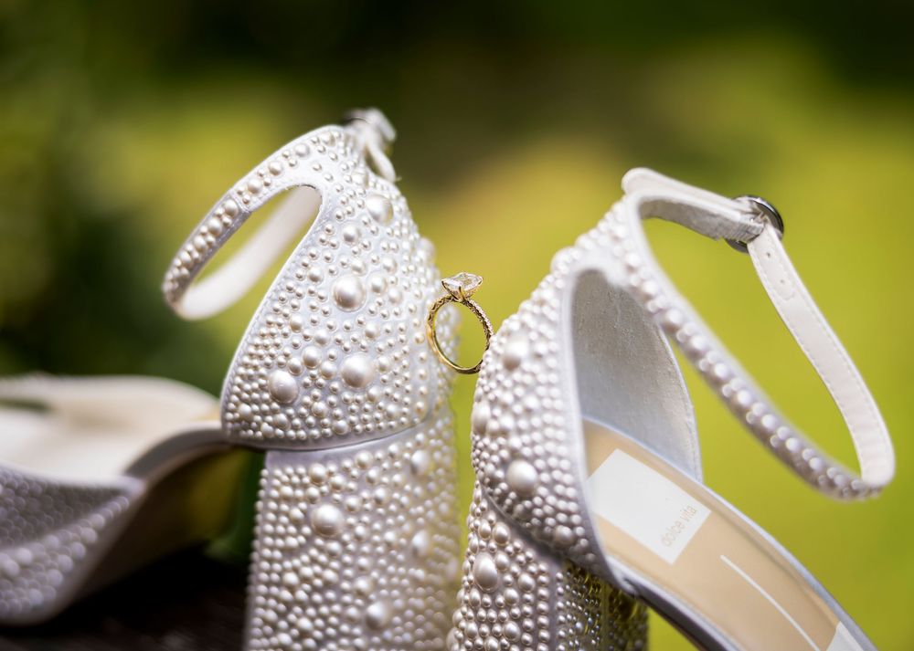 Pearl-encrusted bridal heels with ankle strap showcasing a diamond engagement ring, set against a soft green background.